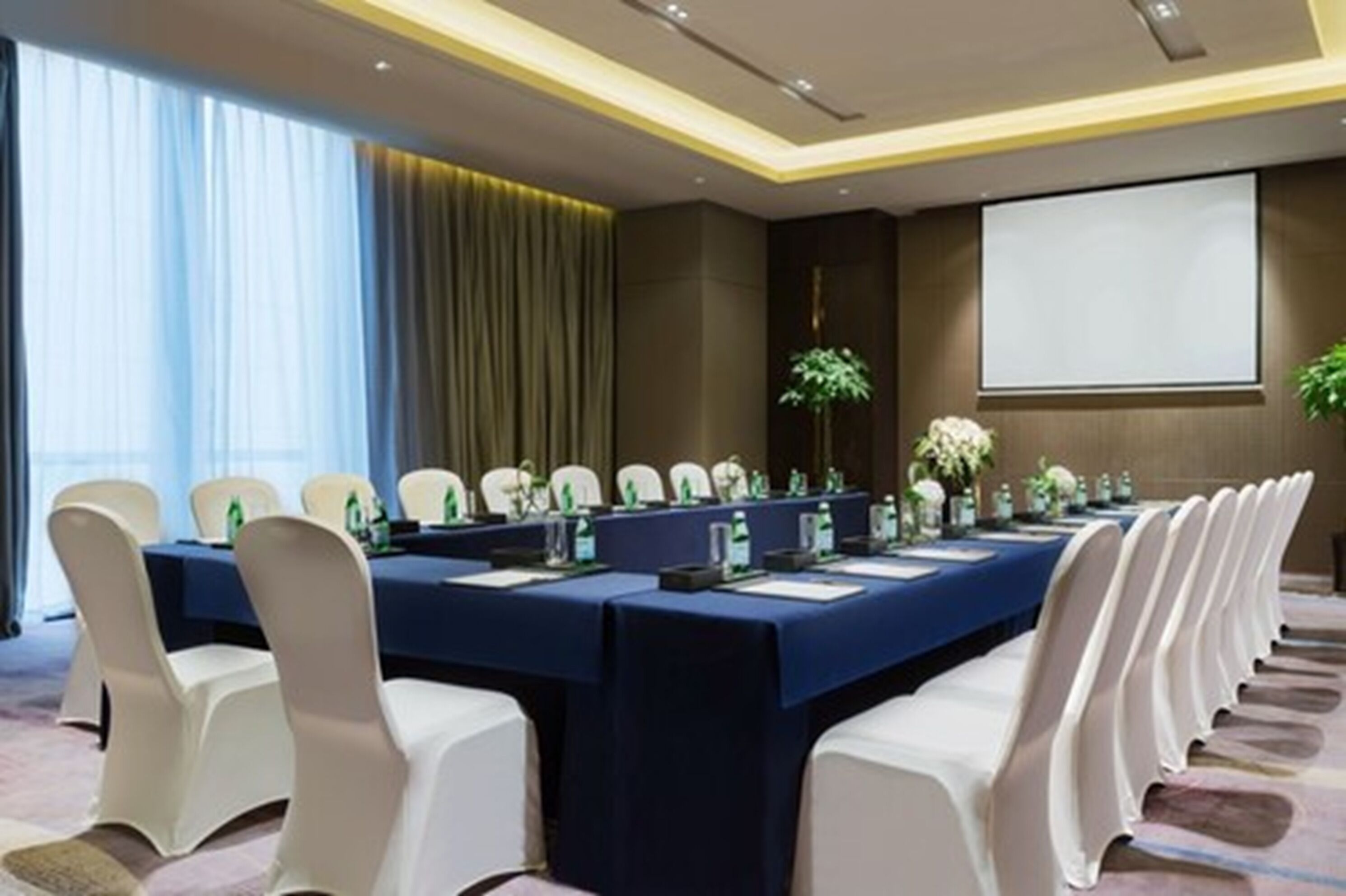 A conference room with a long table, white chairs, and a projector screen.