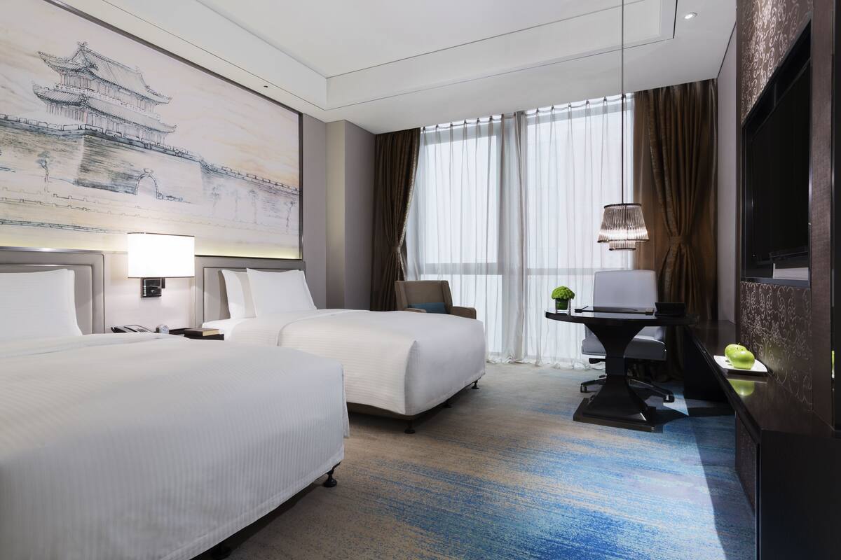 A hotel room with two beds, a desk, a chair, a TV, and a large window with curtains.