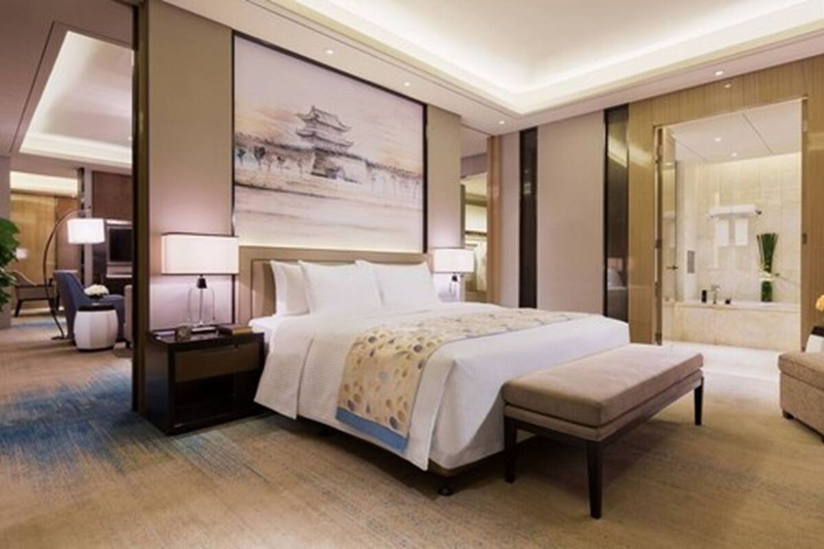 A modern hotel room with a large bed, bedside tables, a nightstand, a bench, a large painting on the wall, and a bathroom with a bathtub and shower.