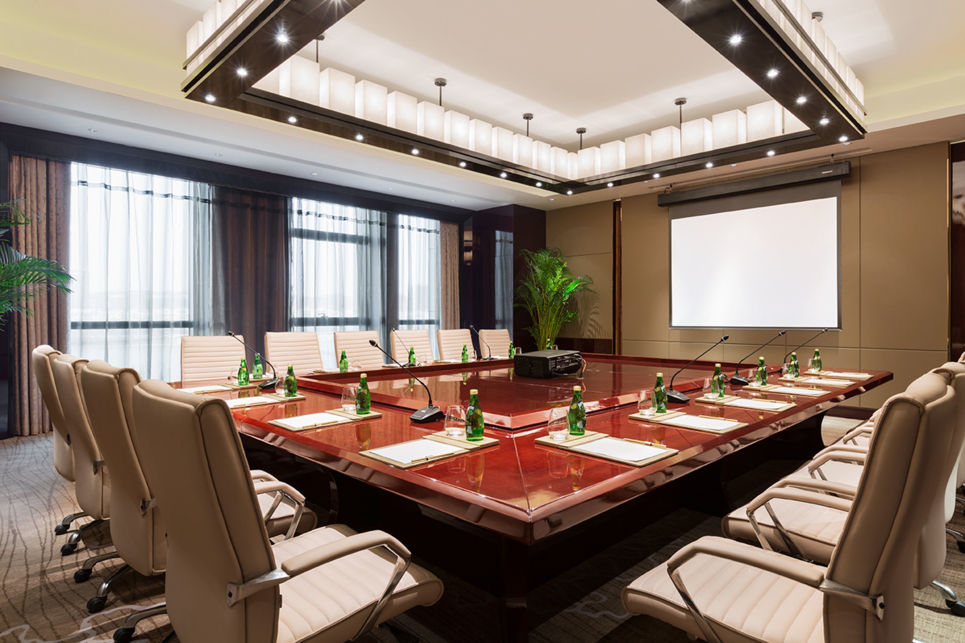 A conference room with a large oval table, beige chairs, a projector screen, and a potted plant.