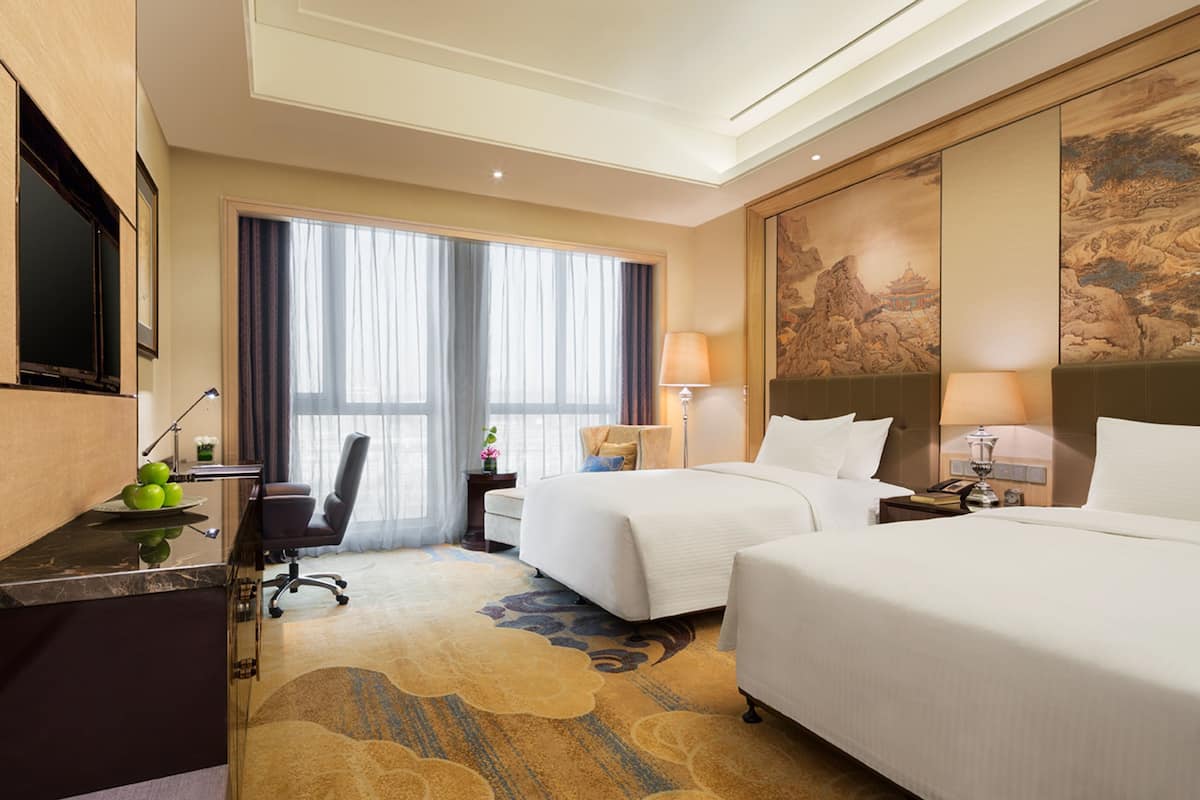 A hotel room with two beds, a desk with a chair, a TV, and a large window with curtains.
