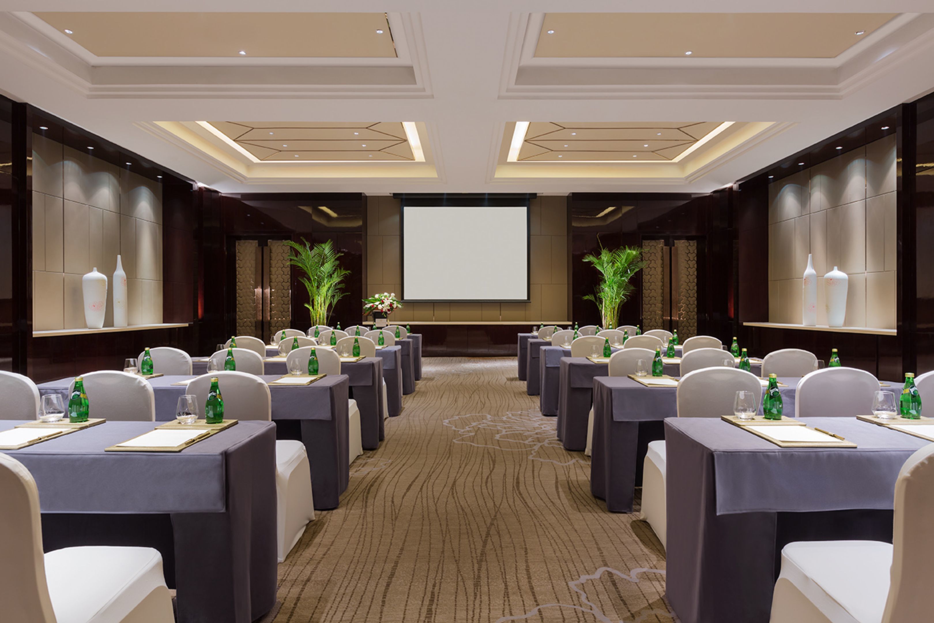 A conference room with rows of tables and chairs, a large screen at the front, and decorative plants.