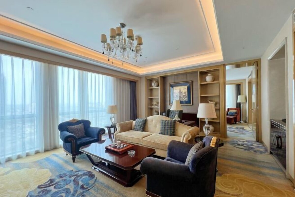 Una habitación de hotel con un ventanal grande, una araña, un sofá, una silla y una mesa de centro.