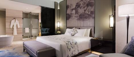 A modern hotel room with a large bed, a sitting area, a bathtub, and a wall-mounted artwork.