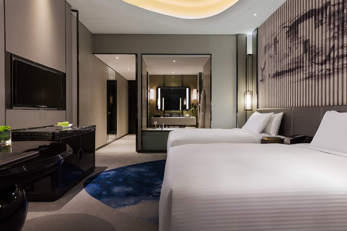 Una habitación de hotel moderna con una cama grande, un escritorio con televisor de pantalla plana, un baño con un espejo amplio y una obra de arte colgada en la pared.