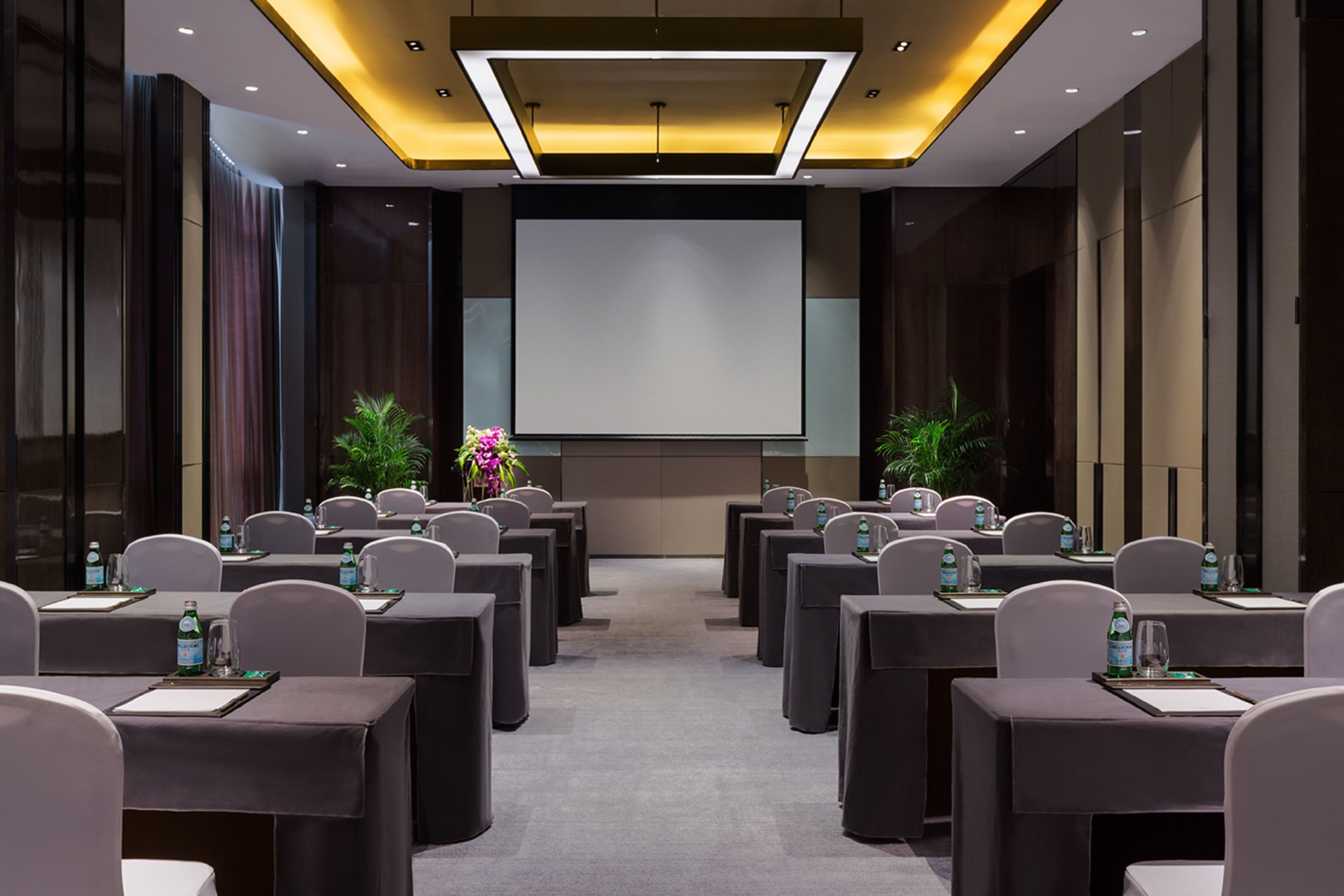 A conference room with rows of tables, chairs, and a large screen at the front.