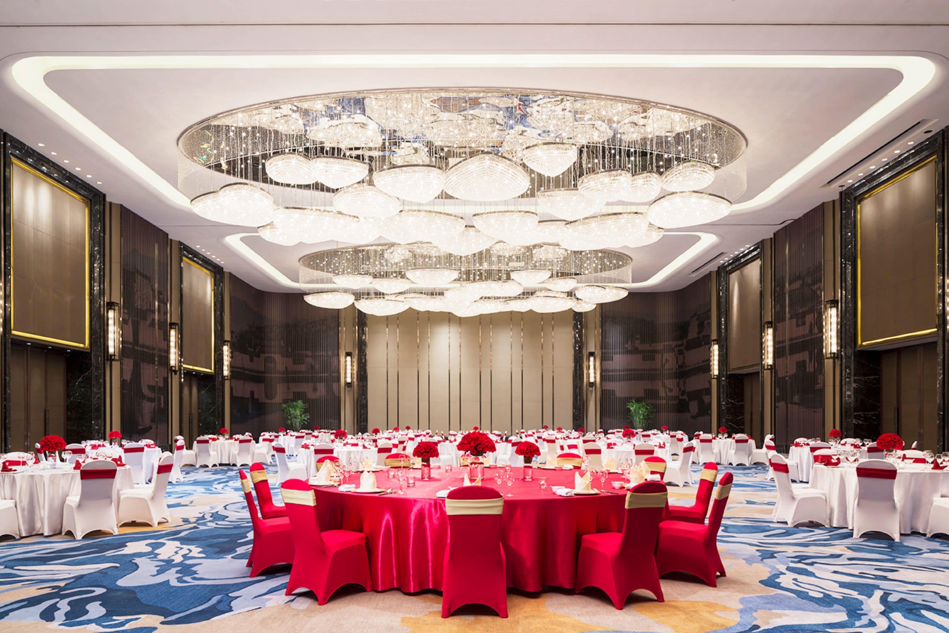 A banquet hall with round tables, red and white tablecloths, and a large chandelier.