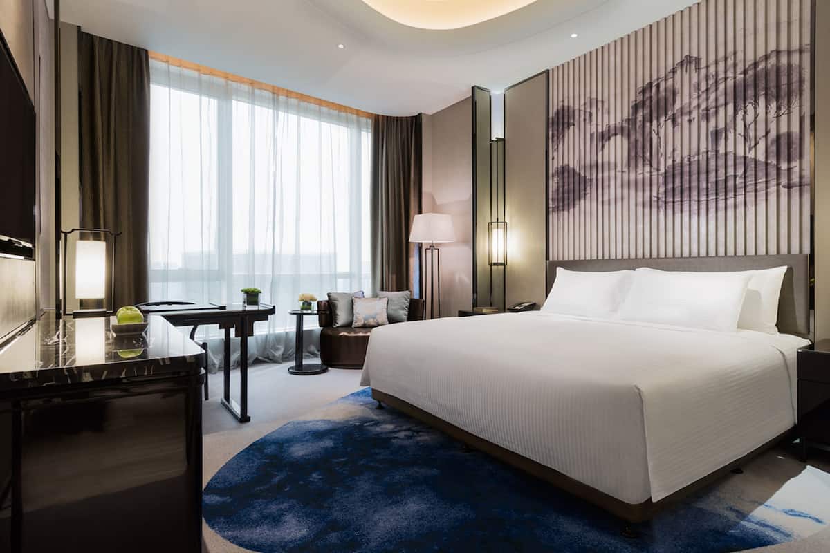 Una habitación de hotel moderna con una cama grande, un escritorio, una silla, una mesita con una lámpara y un ventanal con cortinas.