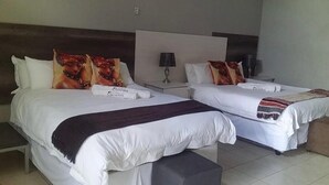 Duas camas de solteiro com roupa de cama branca e almofadas decorativas, uma mesa de cabeceira com abajur e um banco ao pé das camas.