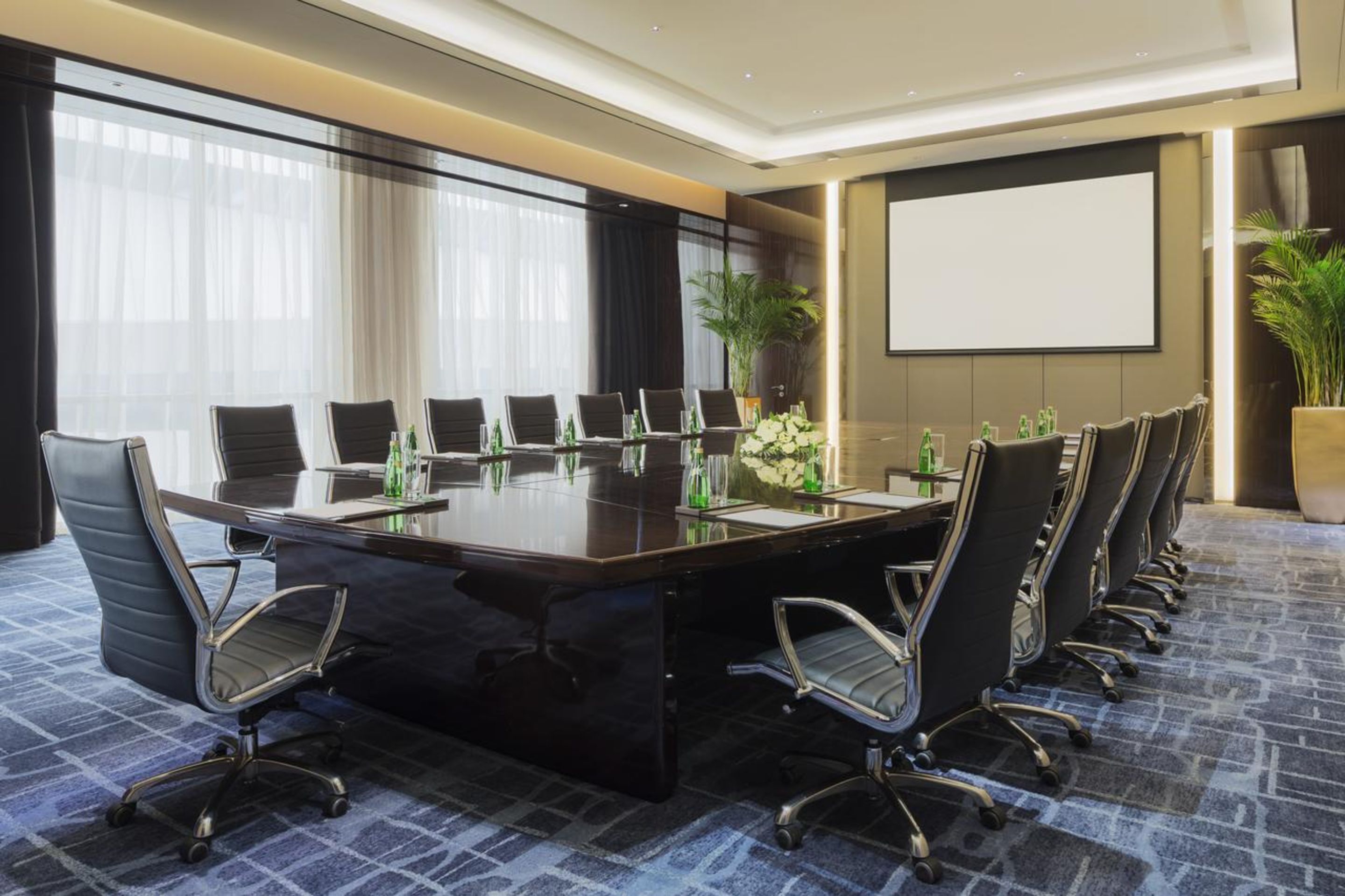 Un salón de conferencias con una larga mesa, varias sillas, una pantalla de proyector y plantas en macetas.