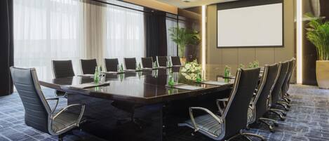 Un salón de conferencias con una larga mesa, varias sillas, una pantalla de proyector y plantas en macetas.