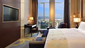 A hotel room with a large bed, a desk, a chair, and a TV. There is a view of a cityscape through the window.