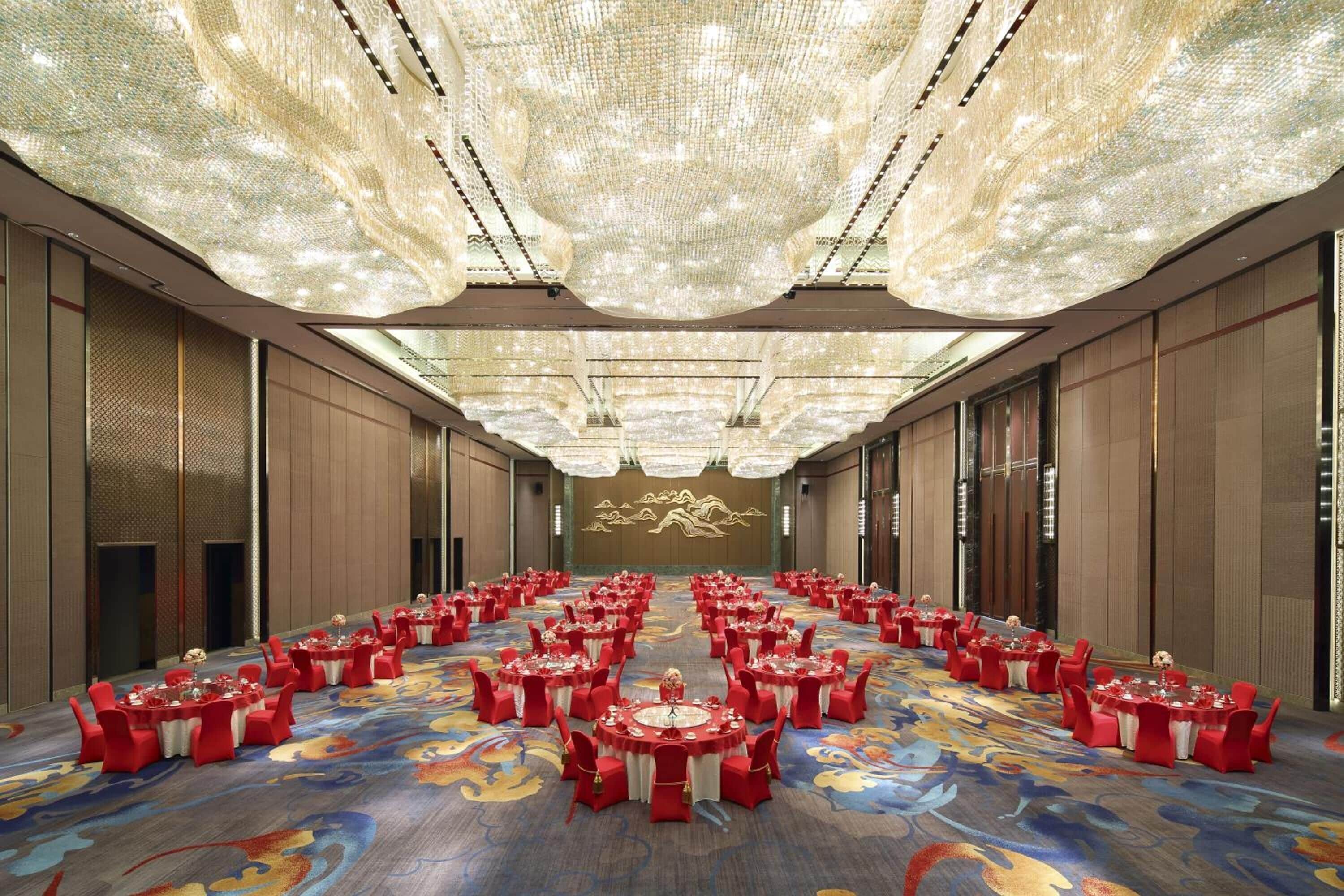 Un salón de banquetes con mesas redondas y sillas rojas, una alfombra estampada y una lámpara de araña grande.