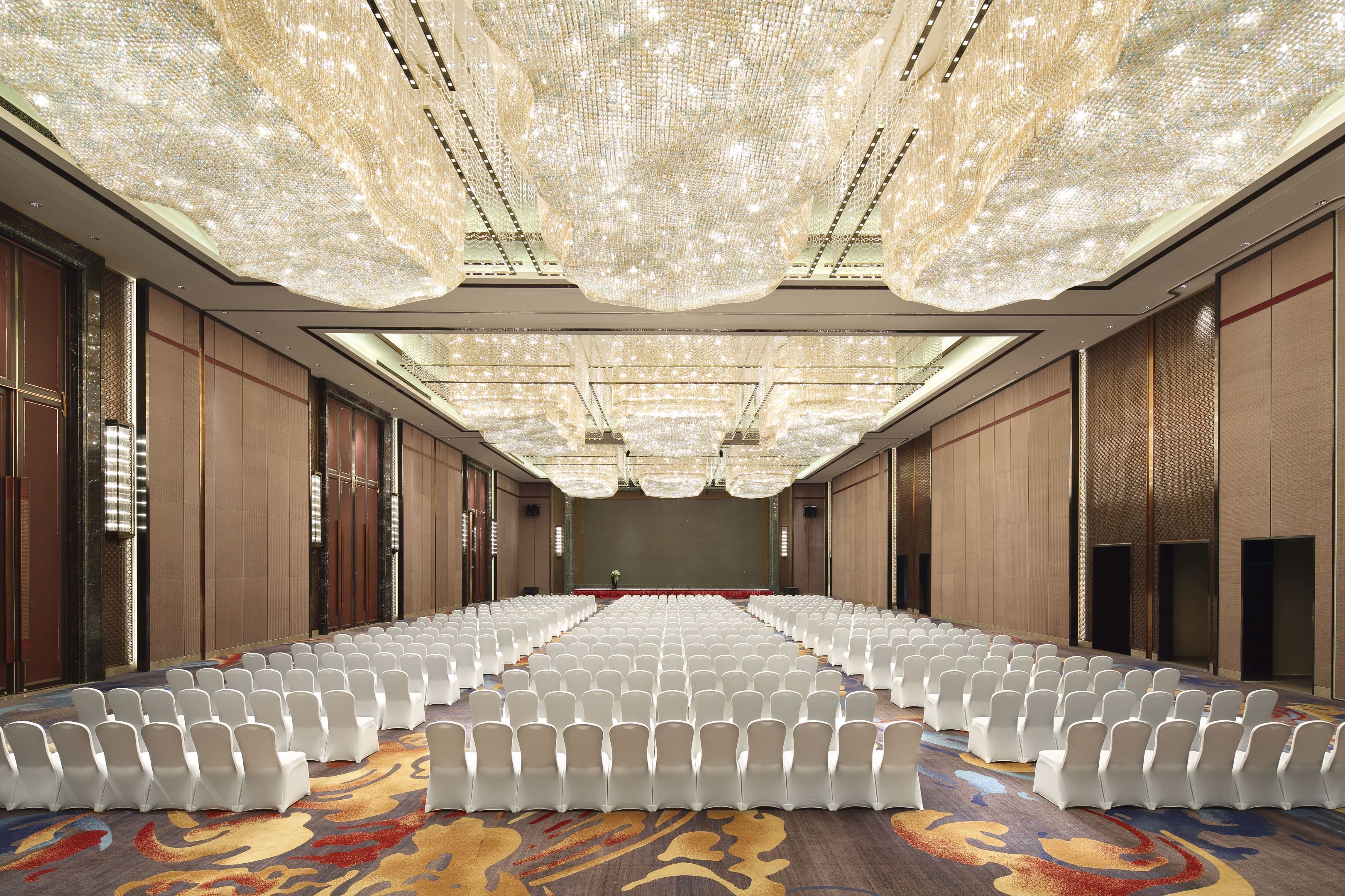 Un'ampia sala conferenze con file di sedie bianche, una moquette decorata e un lampadario.