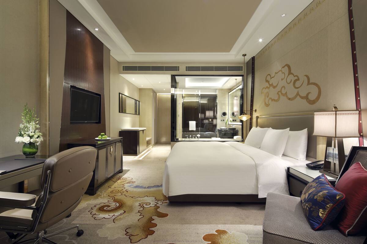 Habitación de hotel con una cama grande, un escritorio con silla, televisor y baño visible a través de una puerta de cristal.