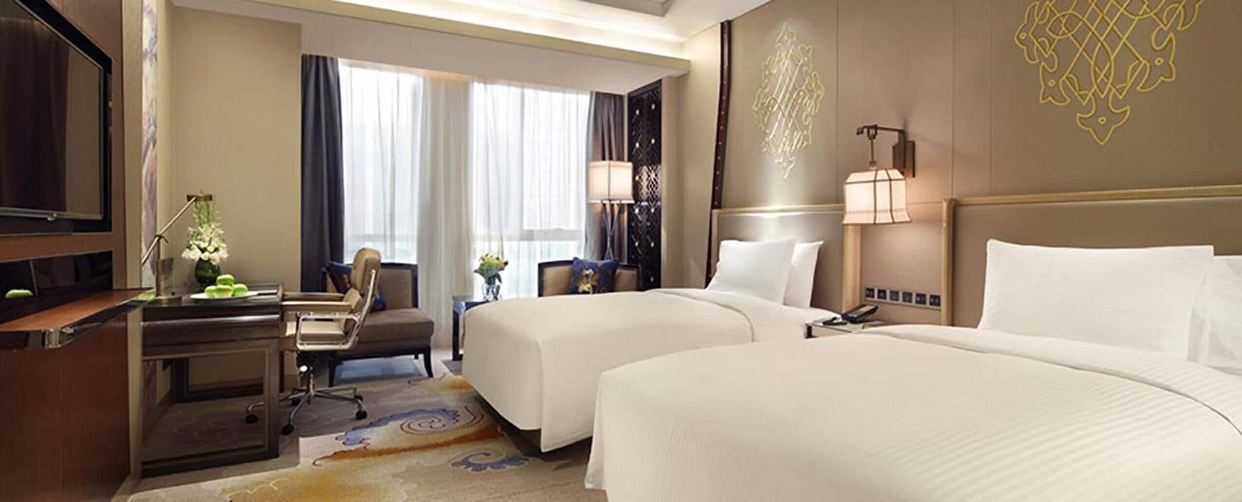 A hotel room with two beds, a desk, a chair, a television, and a window with curtains.