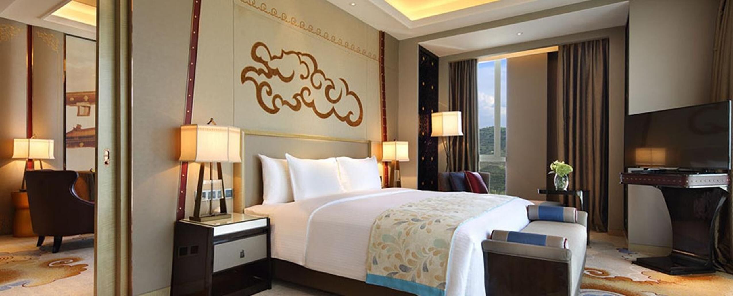 A hotel room with a large bed, two bedside tables with lamps, a TV, and a view of the outdoors.