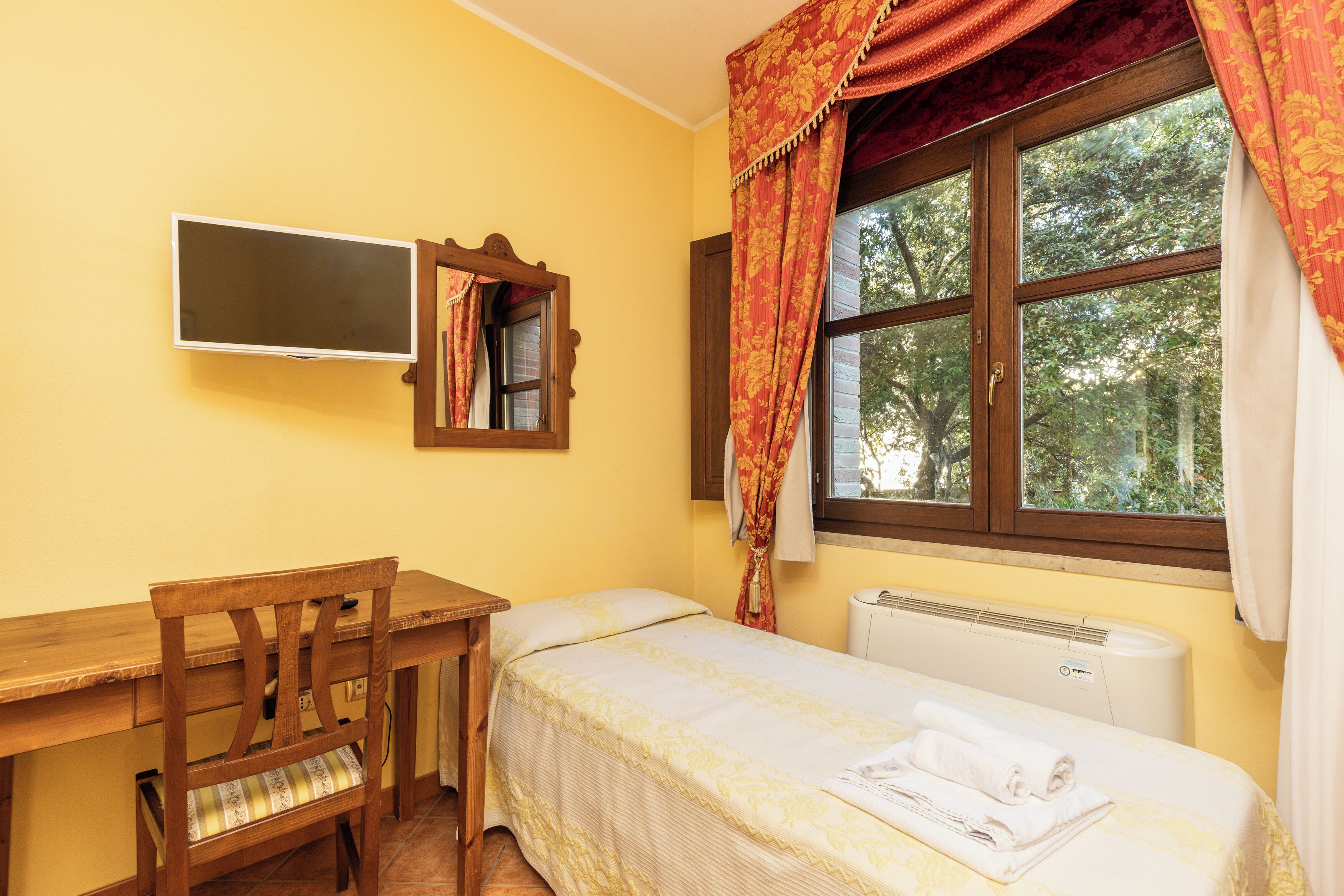 Ein kleiner, ordentlicher Raum mit Schreibtisch aus Holz, einem Einzelbett, einem Heizkörper und einem Fenster mit Vorhängen.