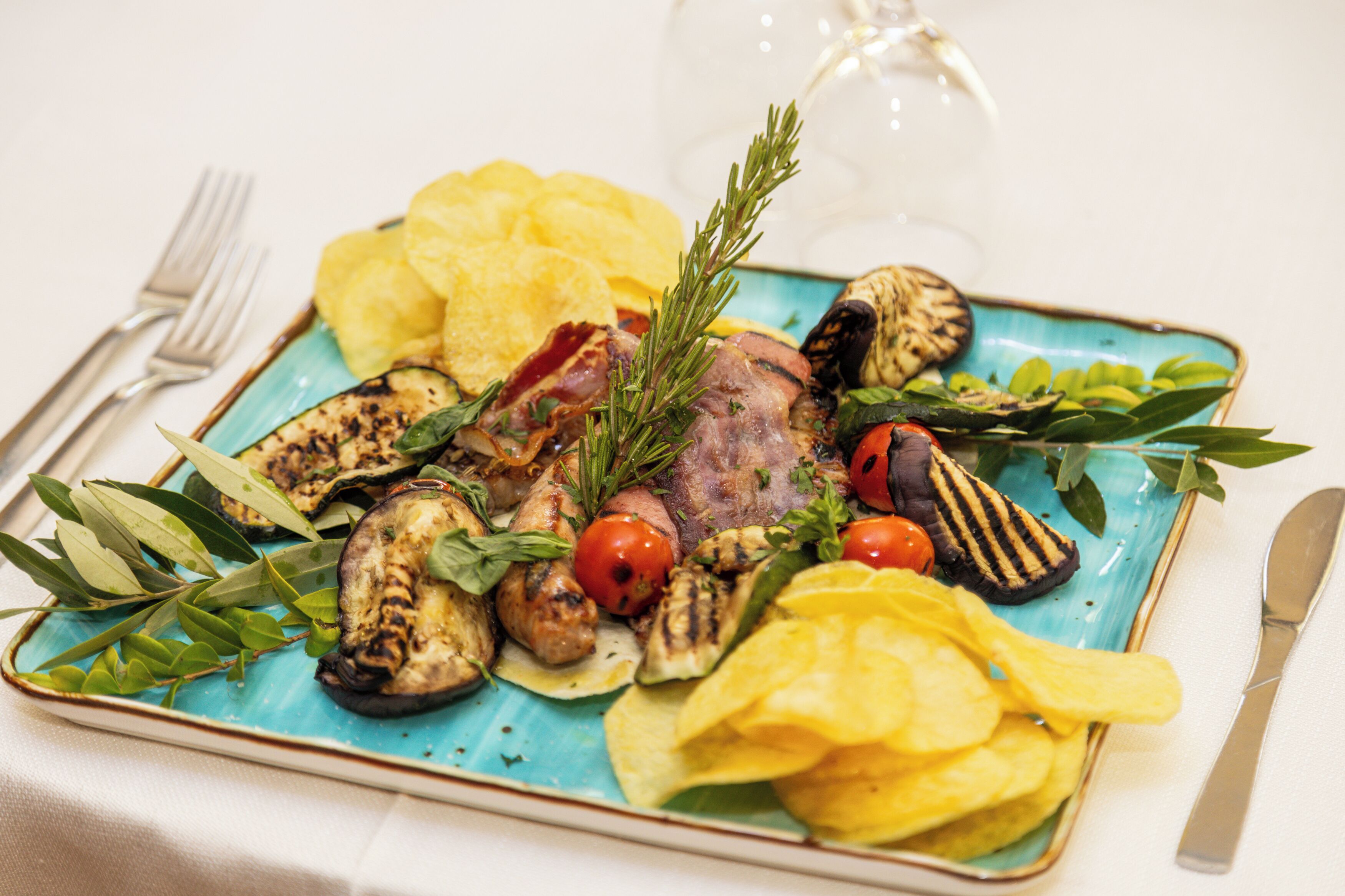 Un plato con verduras a la parrilla, carne y papas fritas sobre una mesa, con un tenedor y un cuchillo.