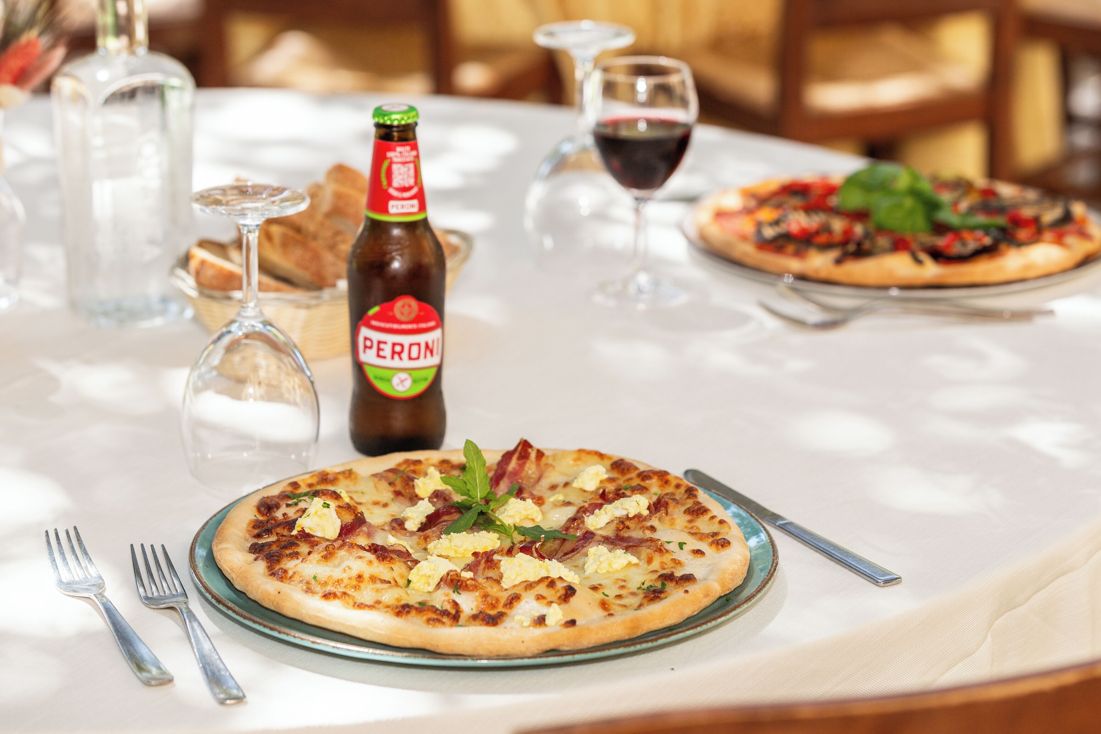 Een eettafel met pizza, een fles Peroni bier, wijnglazen en bestek.