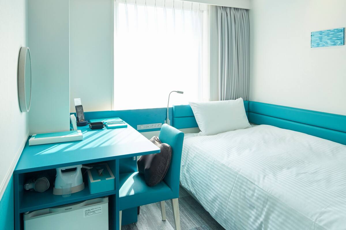 A hotel room with a bed, a desk with a computer, a chair, a small cabinet, and a window with curtains.