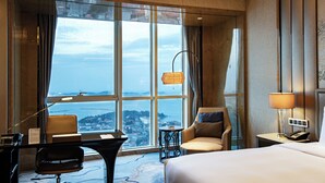 A hotel room with a large window, a desk, a chair, a bed, and a nightstand with a lamp. - Pullman Yantai Center (Yantai)