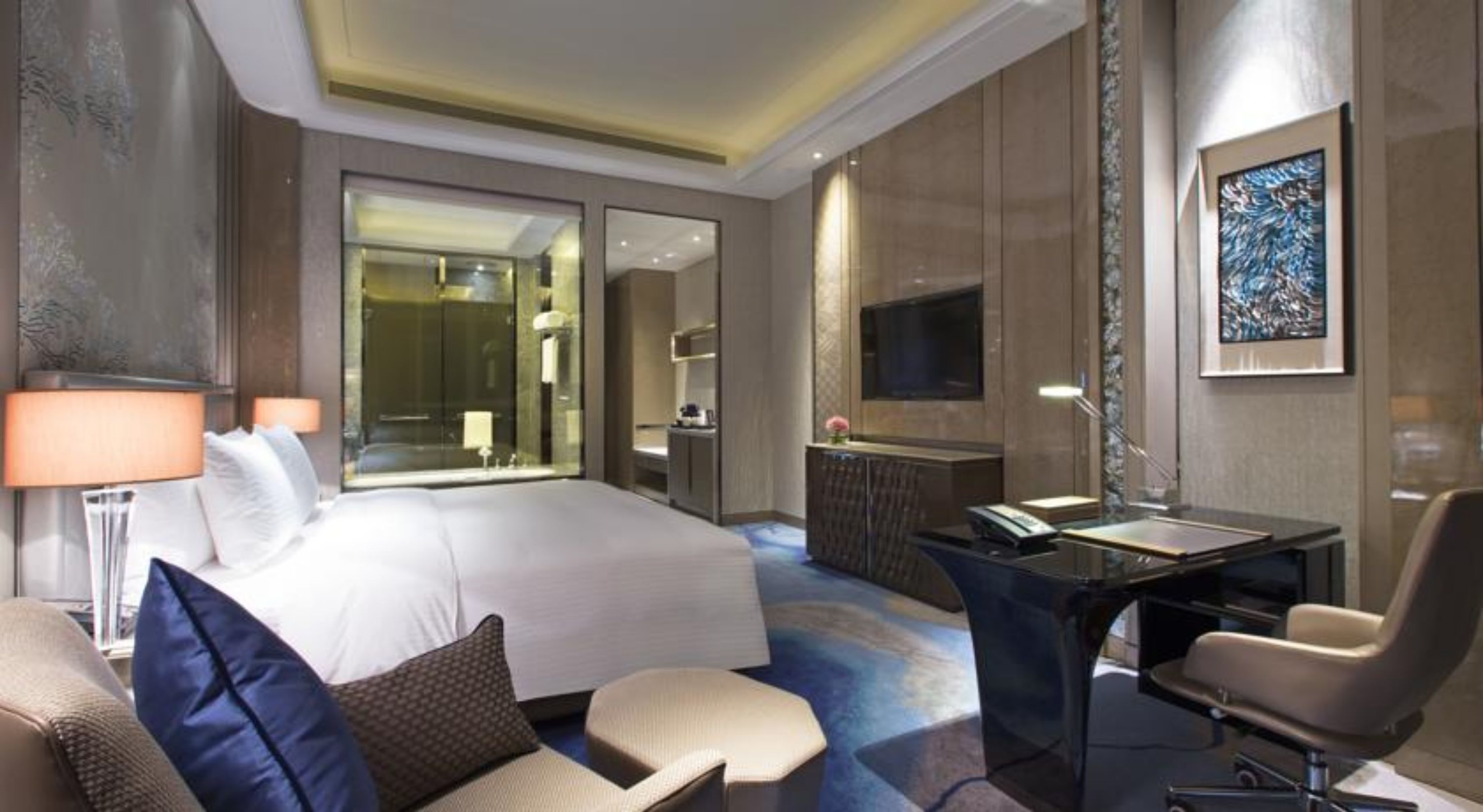 A hotel room with a large bed, a desk, a chair, a TV, and a bathroom visible through a glass door.