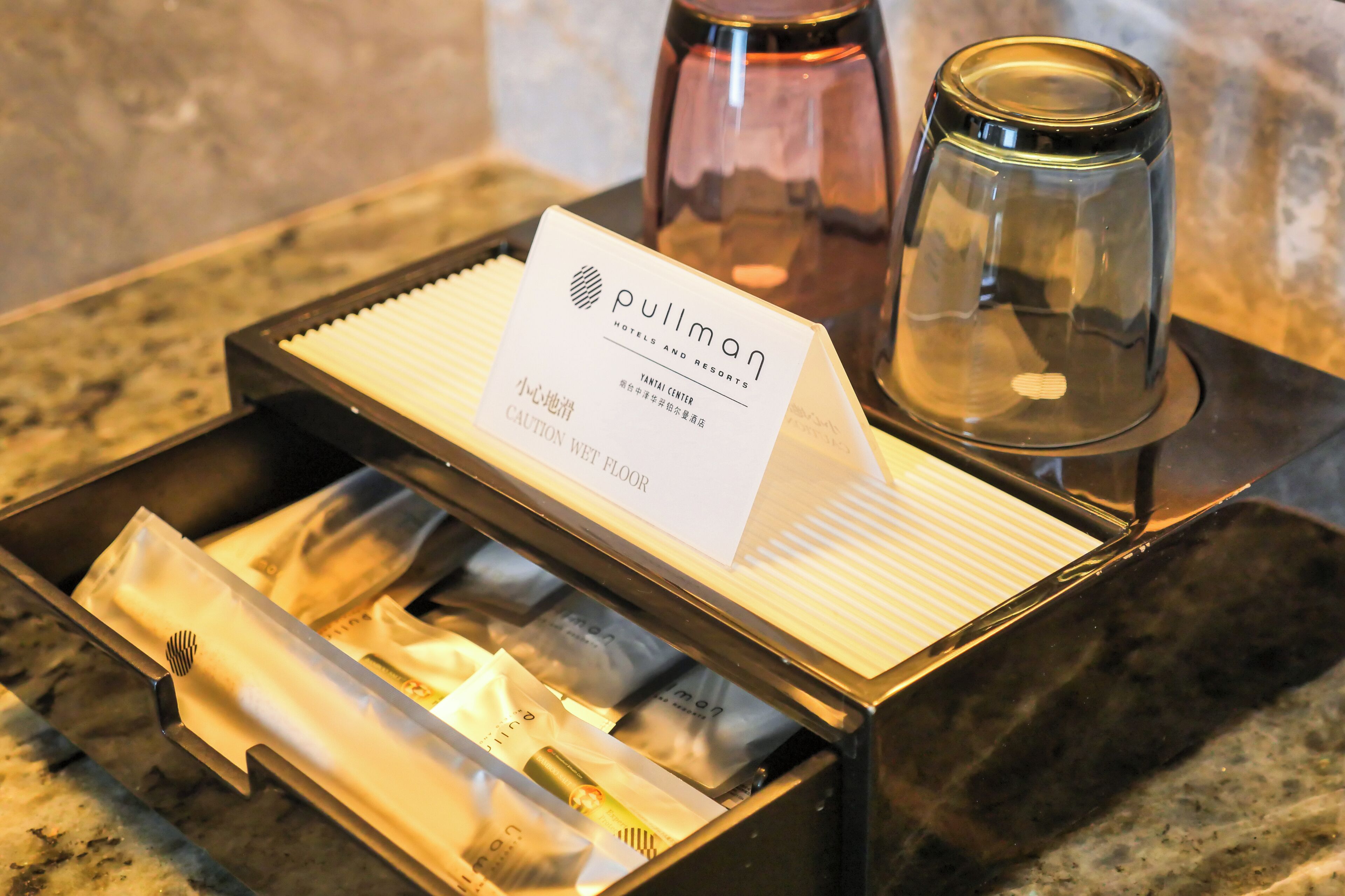 Een hotelminibar met een Pullman-merk kaart, een glazen bak en diverse toiletartikelen.