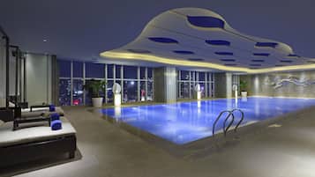 A modern indoor swimming pool with a city view, a lounge area with seating, and a unique ceiling design.