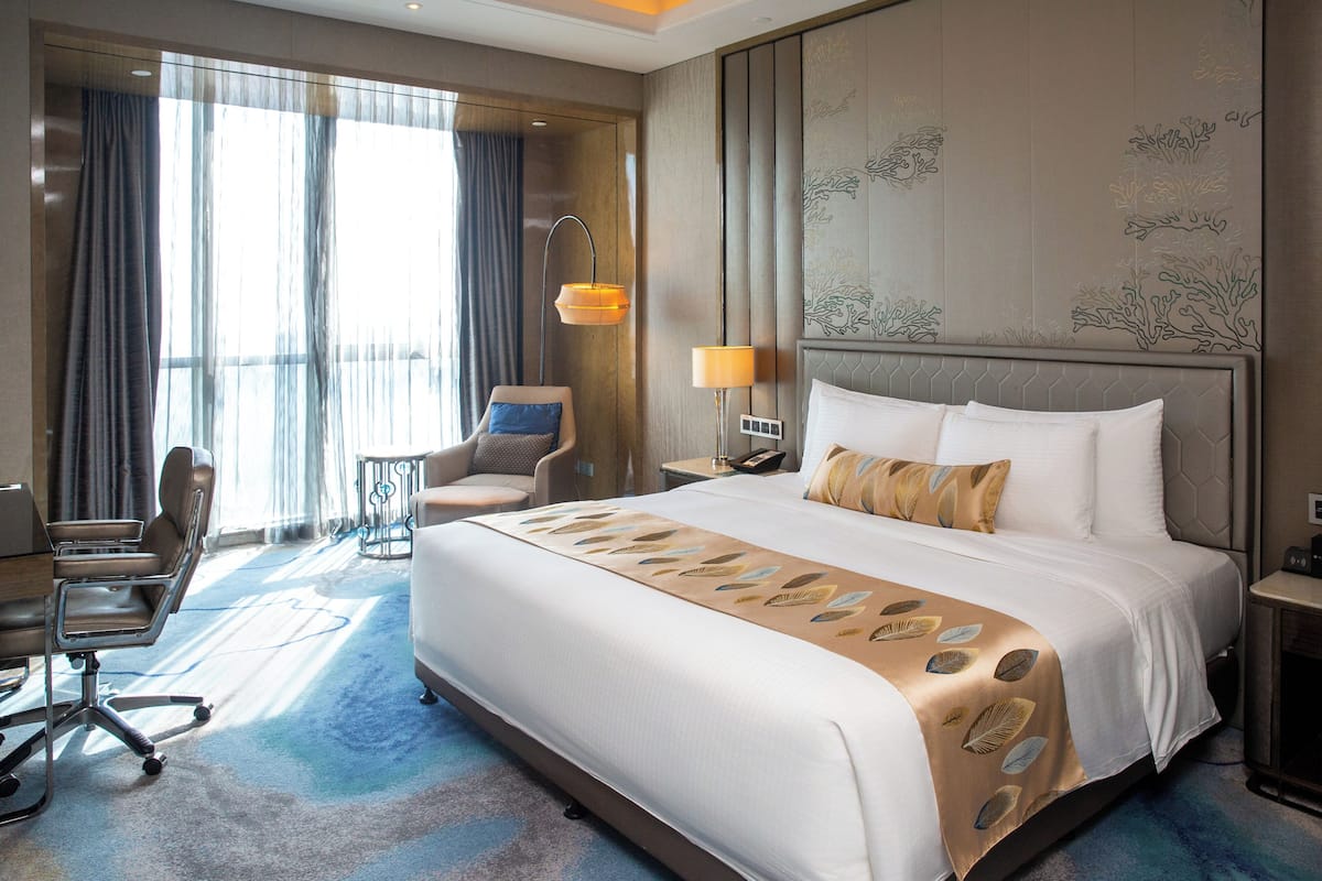 A hotel room with a large bed, a desk, a chair, a lamp, and a view of the outdoors.