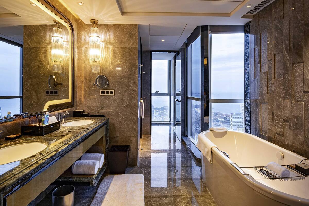 A modern bathroom with two sinks, a large mirror, a chandelier, and a bathtub.
