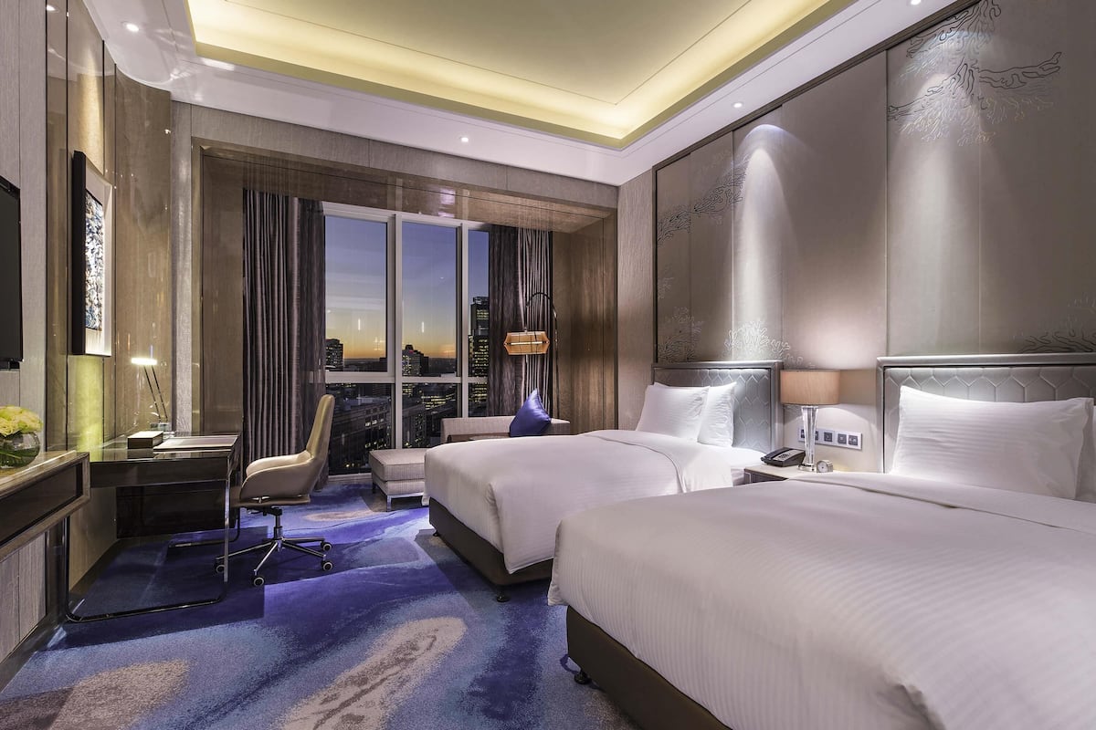 Quarto de hotel com uma cama grande, uma cama menor, uma escrivaninha, uma cadeira e vista para a cidade.