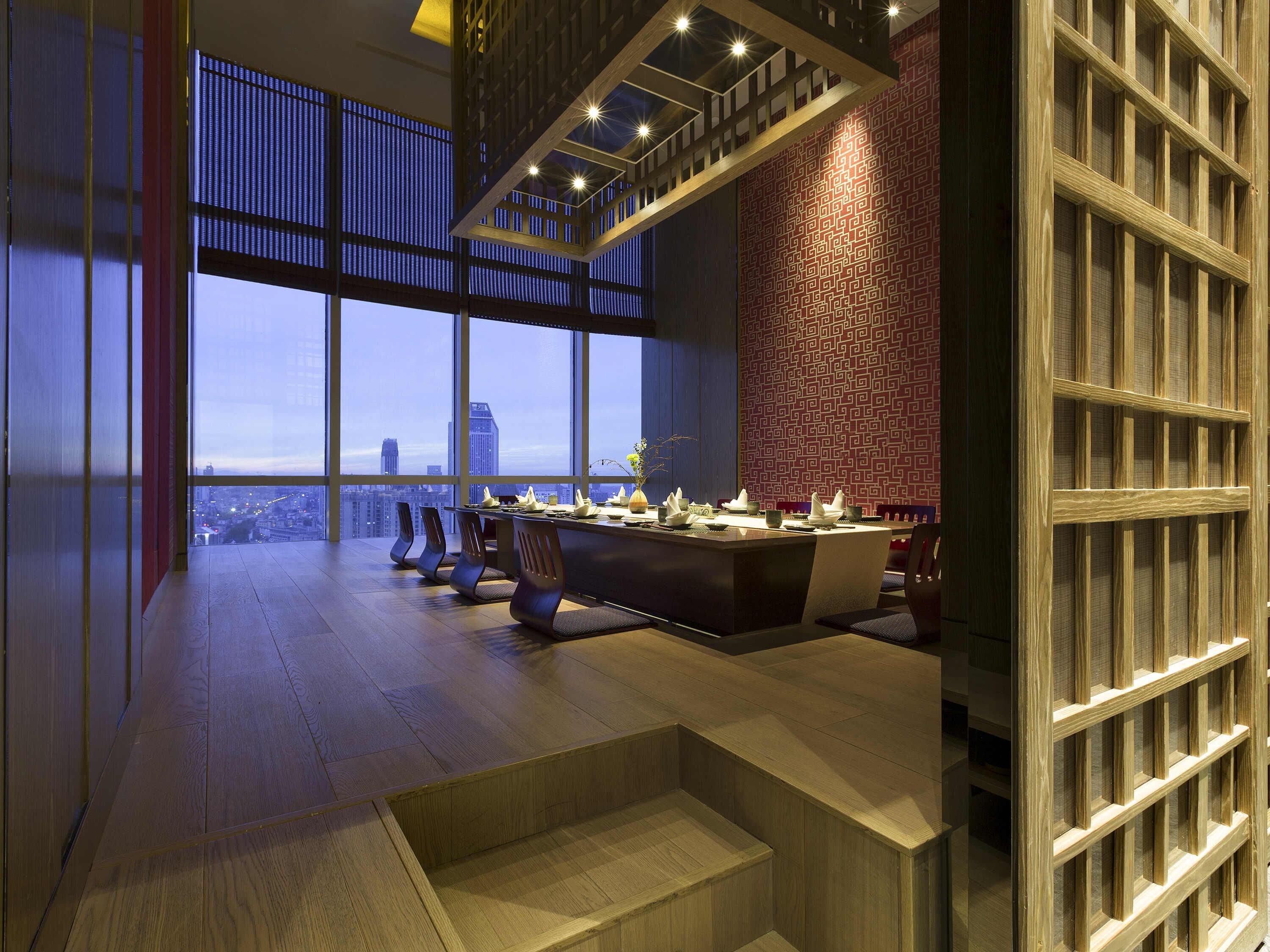 A modern dining area with a high ceiling, large windows, and a city view.