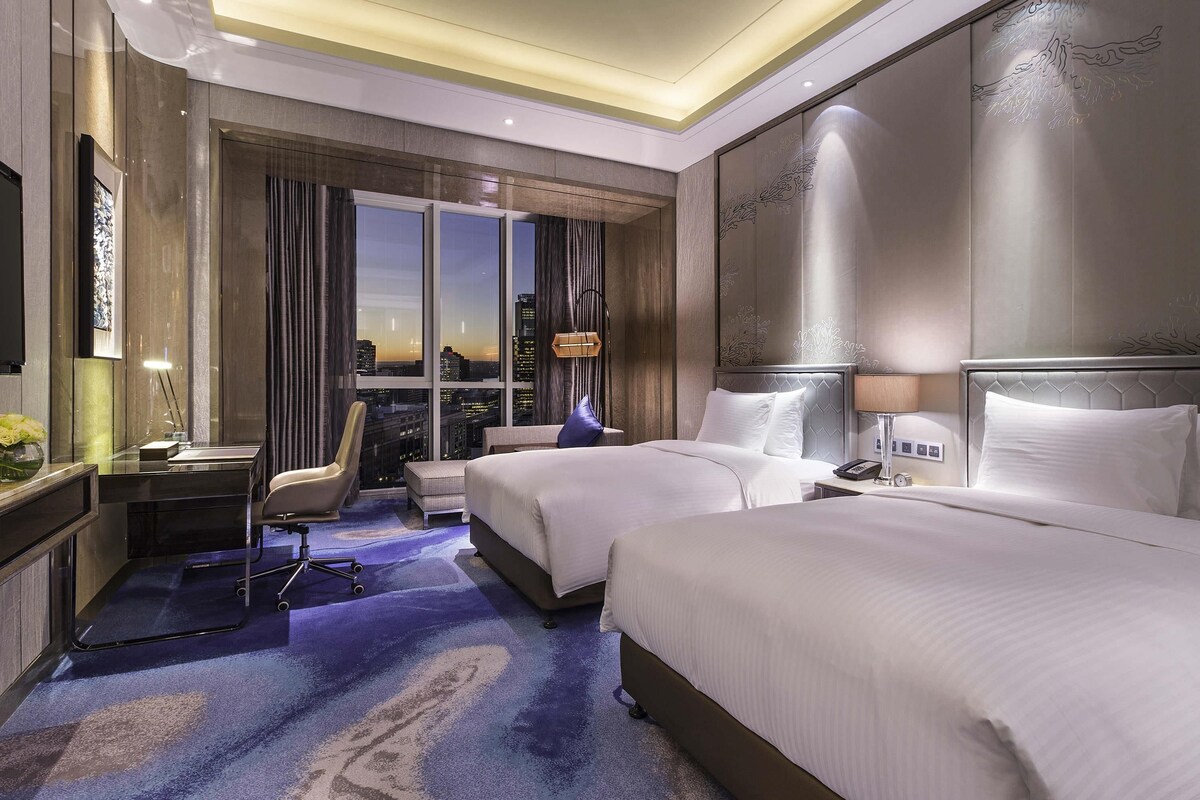 A hotel room with a large bed, a smaller bed, a desk, a chair, and a view of the cityscape.