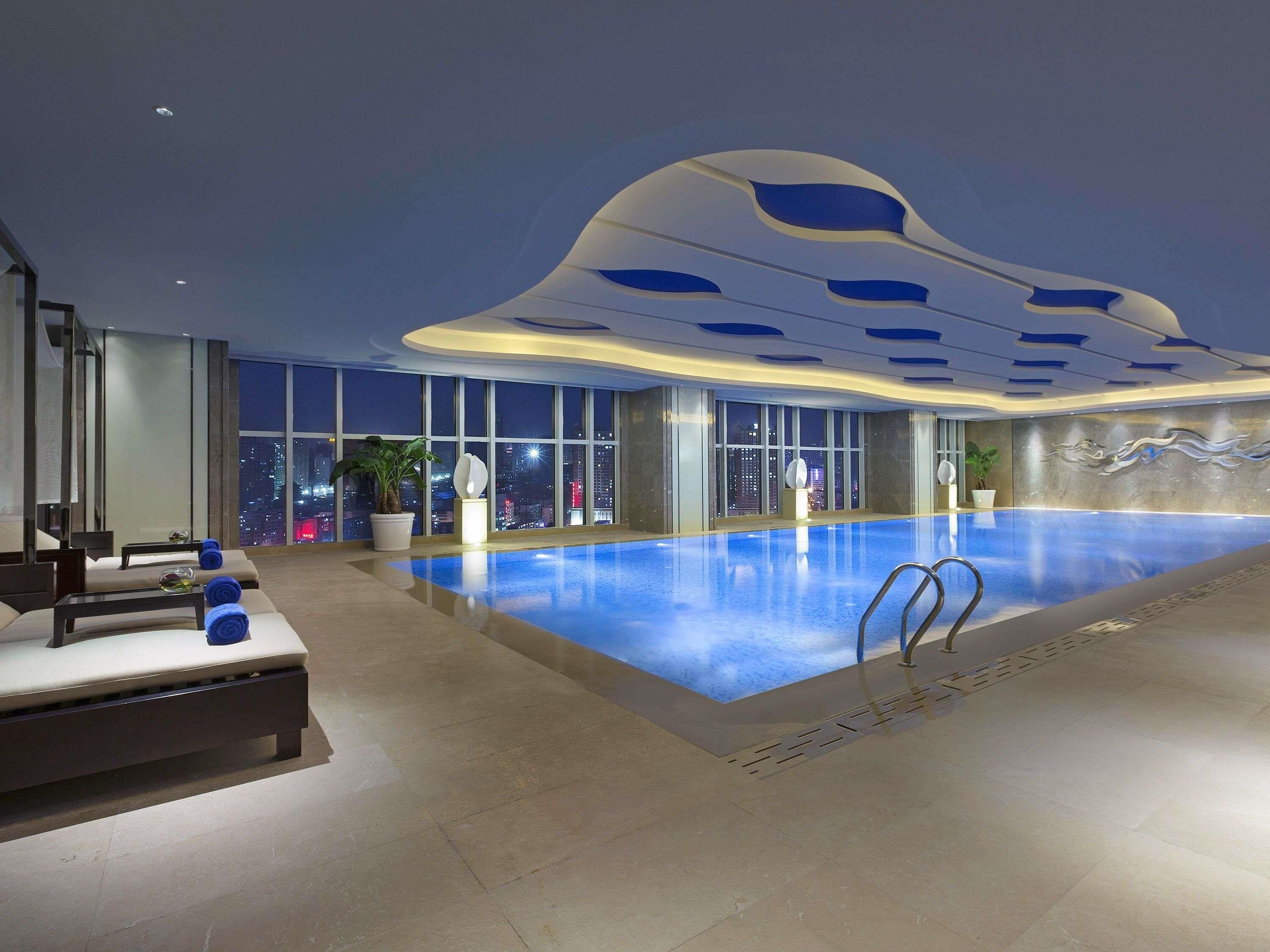Una piscina interior moderna con vista a la ciudad, un área de descanso con asientos y un diseño de techo singular.