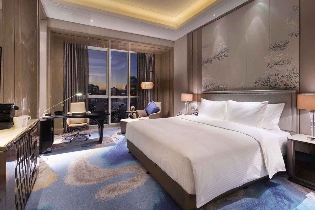 A hotel room with a large bed, a desk with a computer, a chair, and a view of the cityscape through large windows.