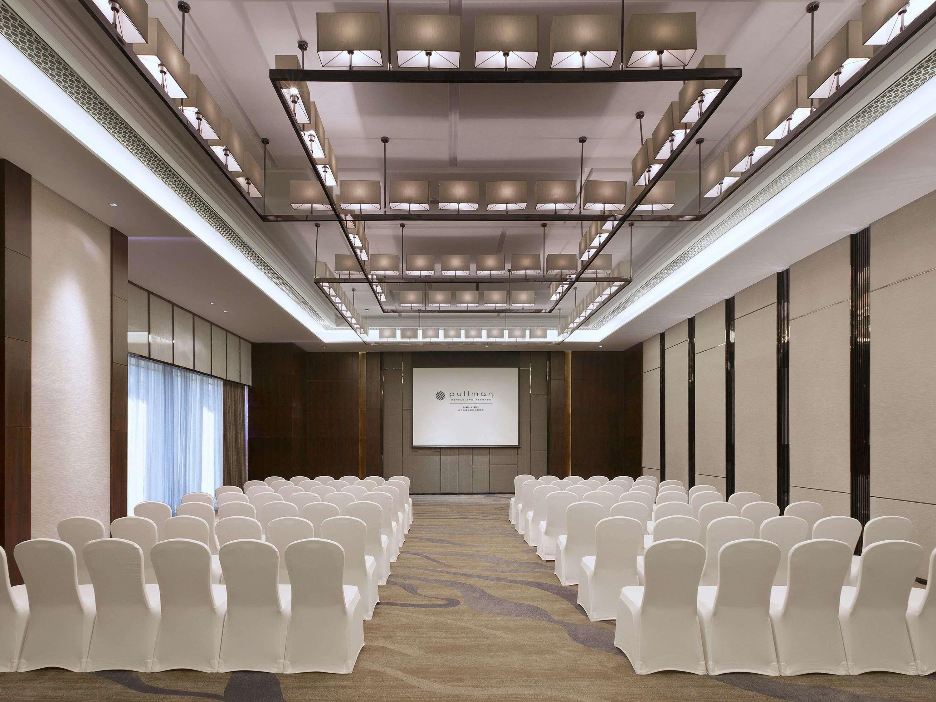 A conference room with rows of white chairs, a projector screen, and a modern ceiling design.