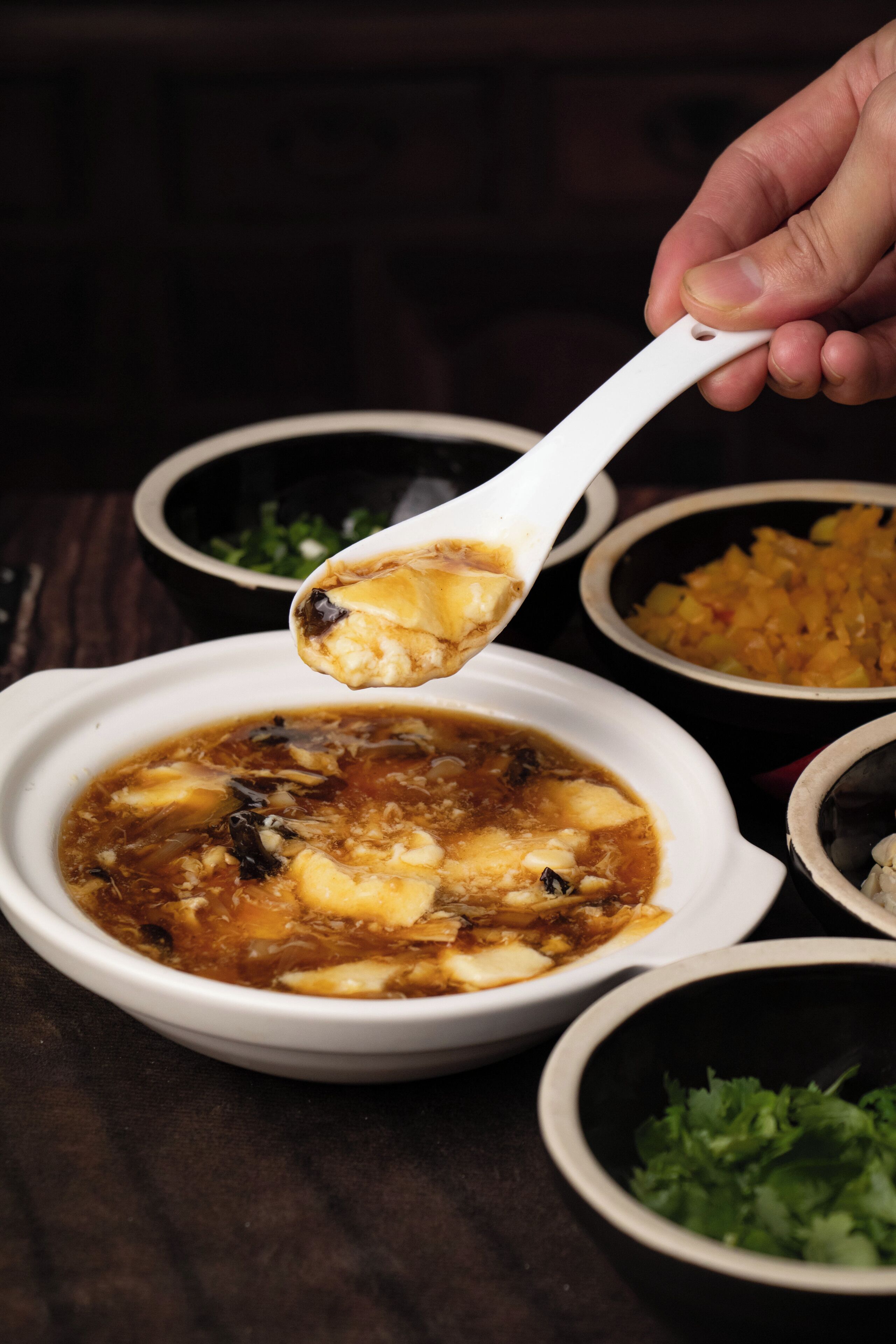 A bowl of soup with a spoon lifting a piece, surrounded by other bowls of food.