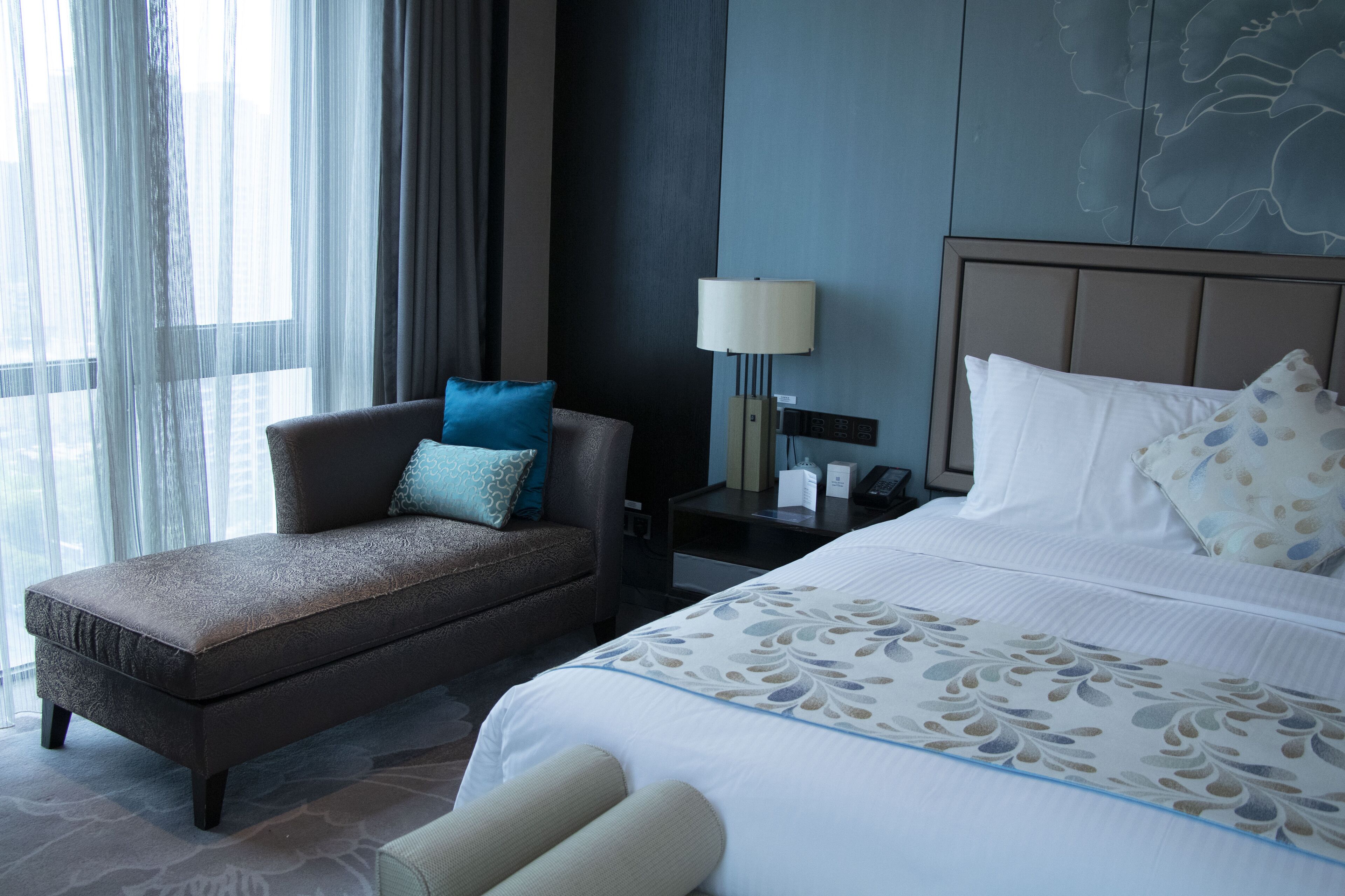 Quarto de hotel com cama, sofá, mesa de cabeceira, luminária e janela com cortinas.