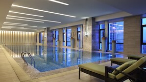 Una piscina interior moderna con agua cristalina, sillones de descanso y grandes ventanales con vistas a la ciudad.