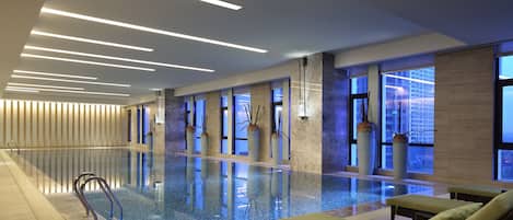 A modern indoor swimming pool with clear water, lounge chairs, and large windows offering city views.