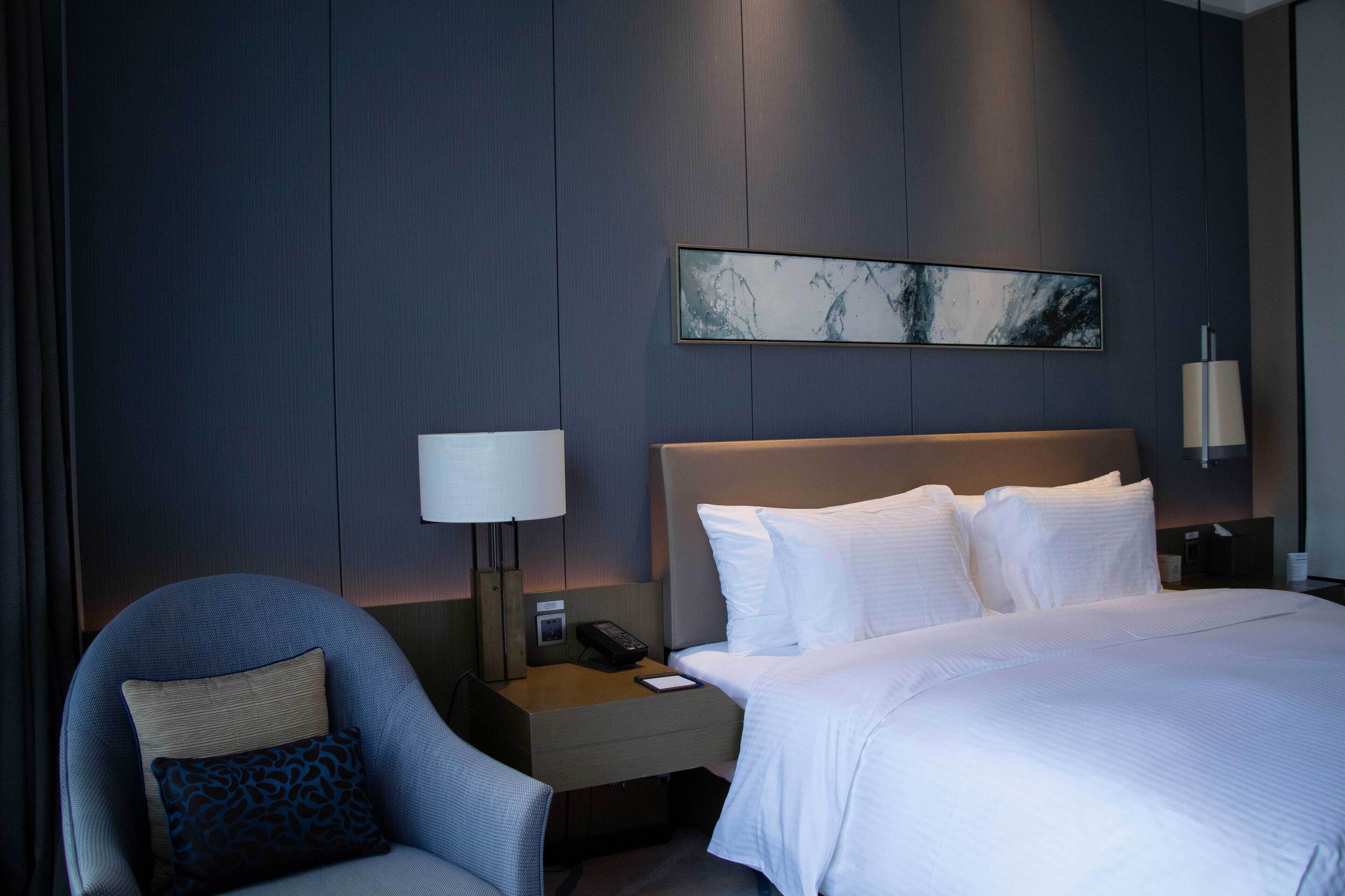 A hotel room with a bed, a chair, a nightstand with a lamp, and a wall-mounted artwork.