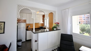 Cocina compacta con armarios blancos, un refrigerador, un microondas y un lavamanos. Hay una ventana con vistas a edificios y una planta con flores en la encimera.