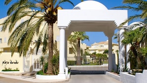 Entrada de hotel con un arco blanco, palmeras y un cielo azul despejado.