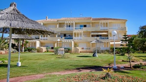 Un hotel de varios pisos con fachada amarilla, balcones y un jardín bien cuidado con un toldo de paja.