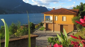 Une maison à deux étages dotée d’un balcon, d’une allée pavée et offrant une vue sur un lac, avec des montagnes en arrière-plan.
