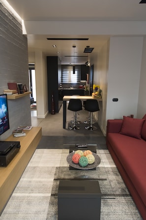 A modern living room with a red sofa, a coffee table with decorative balls, a TV, and a bookshelf. A kitchen is visible in the background with a dining area. - BRAND NEW LUXURY APARTMENT 10' MINUTES WALK FROM ACROPOLIS AND 6' FROM METRO (Athina)