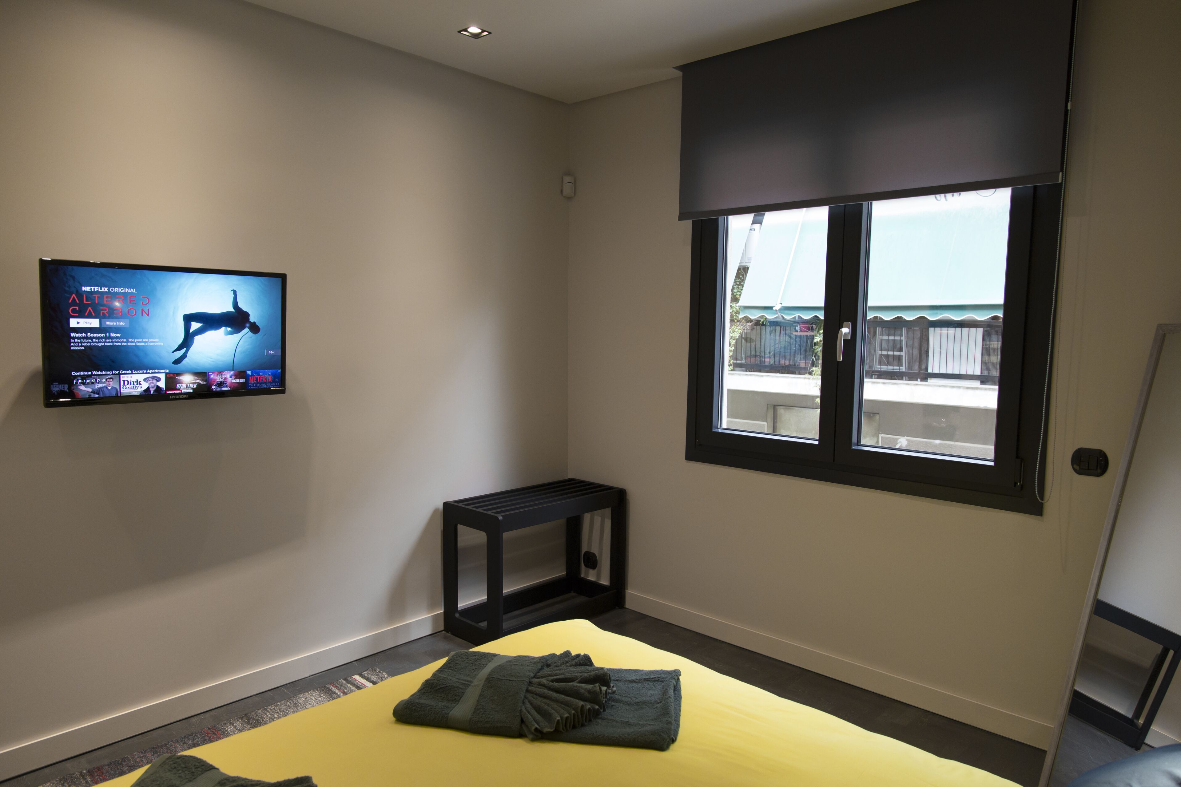 A hotel room with a bed, a TV mounted on the wall, a small table, and a window with a view of the outside.
