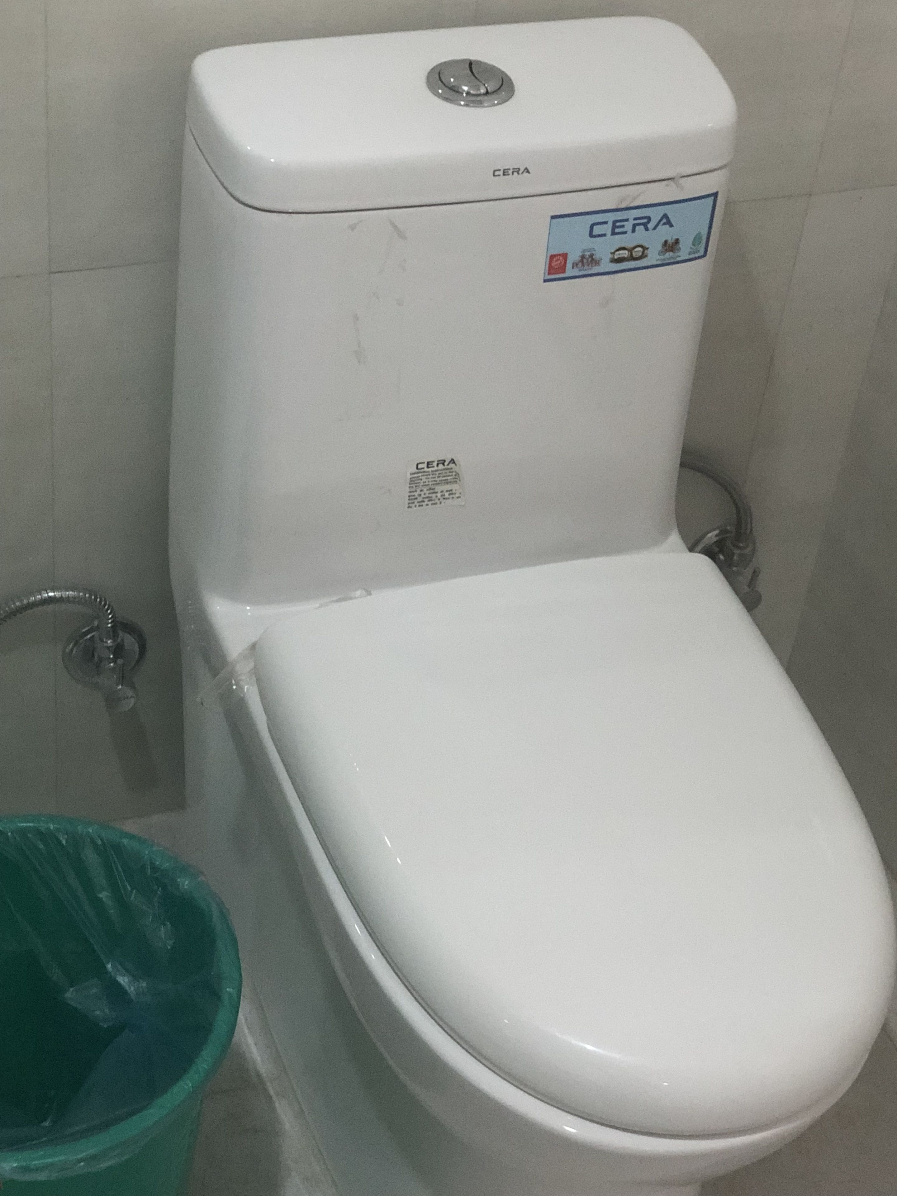 A white toilet with a CERA label, a green trash bin, and a metal pipe.