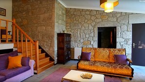 Una sala de estar con pared de piedra, un sofá morado, un sofá estampado, una escalera y un televisor.